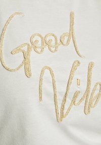 Tessuto bianco con ricamo in filo dorato che forma la scritta "Good Vibes". Le lettere hanno una texture liscia, leggermente in rilievo e un design informale e fluido.