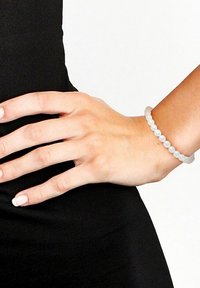 Bracelet en perles avec des perles blanches givrées, port�é sur une main reposant contre un fond en tissu noir. Forme circulaire, texture lisse.