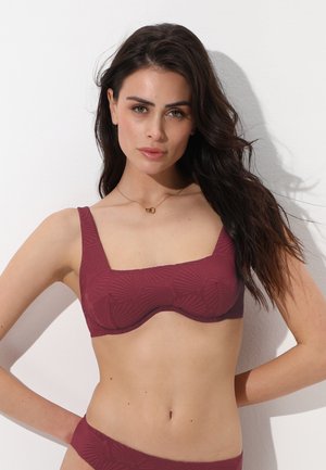 Donna con capelli lunghi e scuri che indossa un top bikini e slip bordeaux testurizzati, in piedi con una mano sul fianco contro uno sfondo bianco.