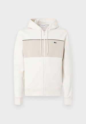 Crème zip-up hoodie met een beige paneel op de borst, verstelbare koordjes, kangoeroe-zakken en een subtiele logo aan de linkerkant.