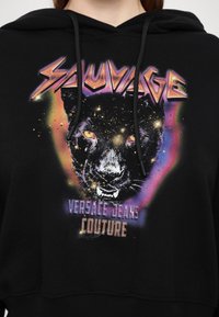 Felpa nera con cappuccio caratterizzata da una stampa grafica del volto di una pantera circondato da dettagli cosmici colorati. Il testo recita "Sauvage" e "Versace Jeans Couture."