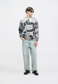 Sweatshirt tie-dye em tons de cinza e creme, combinado com jeans soltos azul claro. O modelo usa óculos de sol pretos e calçado preto.