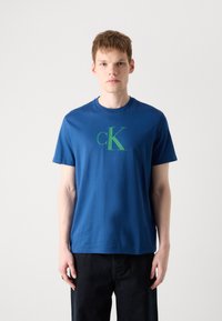 Plava pamučna majica s okruglim izrezom i kratkim rukavima; na prednjem dijelu se ističe istaknuti zeleni logotip "cK"; glatka tekstura.
