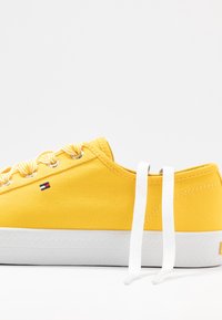 Żółty tenisówka z płótna z białą gumową podeszwą, wyposażona w białe sznurówki i paskowane akcenty. Małe logo umieszczone po boku.