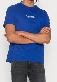 Niebieska bawełniana koszulka z krótkim rękawem, okrągłym dekoltem oraz białym logo "Calvin Klein" nadrukowanym na piersi. Regularny krój, gładka faktura.