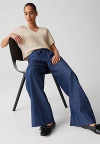 Light beige knitted v-neck sweater with short sleeves paired with loose-fitting dark blue denim trousers. Black flats with bows on feet.