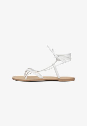 Sandali piatti bianchi presentano fasce sottili, un design minimalista e una suola piatta color beige. Le fasce sono realizzate in un materiale liscio con un dettaglio con lacci.