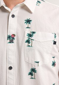 Camicetta bianca a maniche corte, con tessuto testurizzato. Presenta stampe di palme verdi e una tasca a toppa. Dettaglio dei bottoni neri visibile.