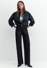 Młoda kobieta stojąca z rękami na biodrach, ubrana w ciemną kurtkę jeansową, biały top, czarne jeansy z wysokim stanem oraz czarne szpilki z ostrym noskiem.