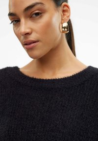 Zwarte gebreide trui met een ronde halslijn, voorzien van een textuurstof. Een gouden ringoorbel voegt een glanzend accent toe aan de algehele look.