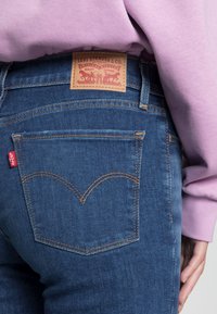 Jeans de mezclilla azul oscuro con un diseño clásico de cinco bolsillos. Parche de cuero marrón en la cintura con logotipo en relieve. Costuras amarillas que resaltan la forma de los bolsillos.