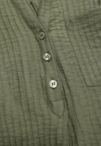 Olijfgroene gestructureerde blouse met een verticale ribbelpatroon, vijf knopen in een bijpassende kleur en een zakdetail aan de linkerkant.