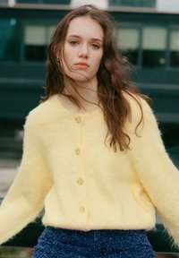 Gelber, flauschiger Cardigan mit rundem Ausschnitt, der fünf Knöpfe auf der Vorderseite hat. Das Model trägt ihn über strukturierten blauen Hosen.