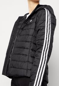 Svart puffjacka med dragkedja, med kviltad textur, vit tre-stripesdesign på ärmarna och en liten logotyp på bröstet.