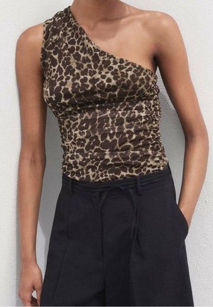 Kvinde iført en one-shoulder top med leopardmønster og sorte højttaljede bukser med hænderne i lommerne mod en hvid væg.