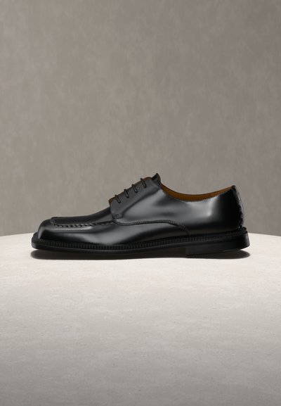 Μαύρο γυαλιστερό δερμάτινο ανδρικό επίσημο παπούτσι με κορδόνια και ραμμένες λεπτομέρειες σε ανοιχτή επιφάνεια με ουδέτερο φόντο.