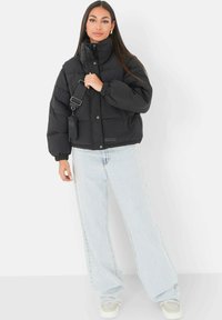 Zwarte gewatteerde jas met hoge kraag, elastische manchetten en drukknoopsluiting, gecombineerd met lichtblauwe wijde jeans en witte sneakers.