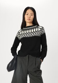 Schwarzer Strickpullover mit einer weißen geometrischen Musterung am Schulterbereich. Lange Ärmel, runder Ausschnitt, kombiniert mit hochgeschnittenen grauen Hosen.