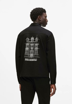 Muž tvárou do strany, ktorý má na sebe čiernu bundu s bielym architektonickým náčrtom a textom „Karl Lagerfeld“ na chrbte na jednoduchom pozadí.