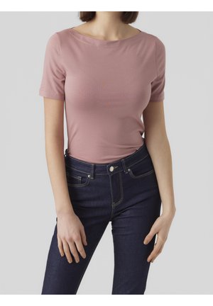 Femme portant un haut rose ajusté à manches courtes rentré dans un jean bleu foncé taille haute, sur un fond gris clair uni.