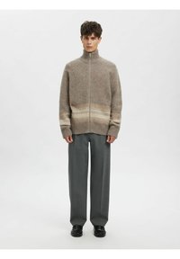 Bēša vilnas džemperis ar rāvējslēdzēju un kontrastējošām horizontālām svītrām. Aprīkots ar augstu apkakli un ribotām aprocēm; kombinēts ar vaļīgām pelēkām biksēm un melniem apaviem.