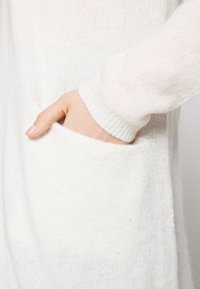 Fehér kötött pulóver laza fazonnal, zsebbel és bordás mandzsettákkal. Puha textúra, mely látható a bőrön.