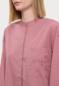 Camicia rosa a colletto realizzata in tessuto morbido, con taschino sul petto dotato di dettaglio ricamato del logo e abbottonatura al collo.