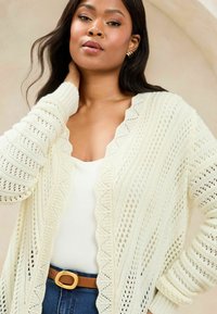 Cremfarbener, gestrickter Cardigan mit gewelltem Saum und offenem Spitzenmuster. Kombiniert mit einem weißen Oberteil und einem braunen Gürtel, vor einem neutralen Hintergrund.