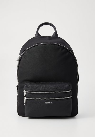 Mochila de tela negra con forma redondeada, que presenta un bolsillo frontal de cuero suave con dos compartimentos con cremallera y acentos de hardware en plata.