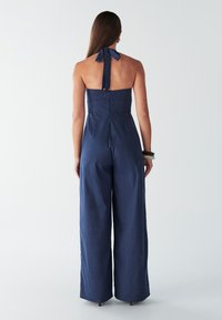 Temno modra kombinezon s halter vrhom in širokimi hlačnicami, zadaj z zavezovanjem in oprijetim zgornjim delom. Izdelan iz teksturirane tkanine, preprost in eleganten dizajn.