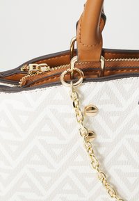 Bolso de mano estampado en beige con asas de cuero color marrón, cremallera dorada y una cadena dorada adjunta cerca de dos tachuelas doradas.