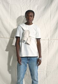 T-shirt bianco di cotone con bordi neri, abbinato a un leggero felpa grigia drappeggiata sulle spalle, indossato con jeans di denim azzurri.