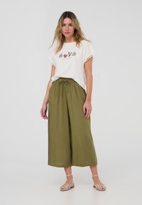 T-shirt bianca con grafica di cactus e bordi in pompon abbinata a pantaloni larghi verde oliva. L'outfit è completato da sandali beige con cinturini.