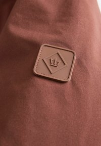 Smeđa tkanina s mat završnom obradom, s kvadratnom smeđom kožnom naljepnicom koja ima utisnutu krunsku oznaku u sredini.