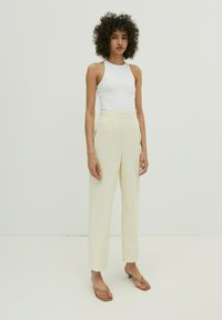 Cremfarbene hochgeschnittene Hose mit geradem Bein und vorderem Nähdetail, kombiniert mit einem engen weißen Tanktop und beigen Sandalen.
