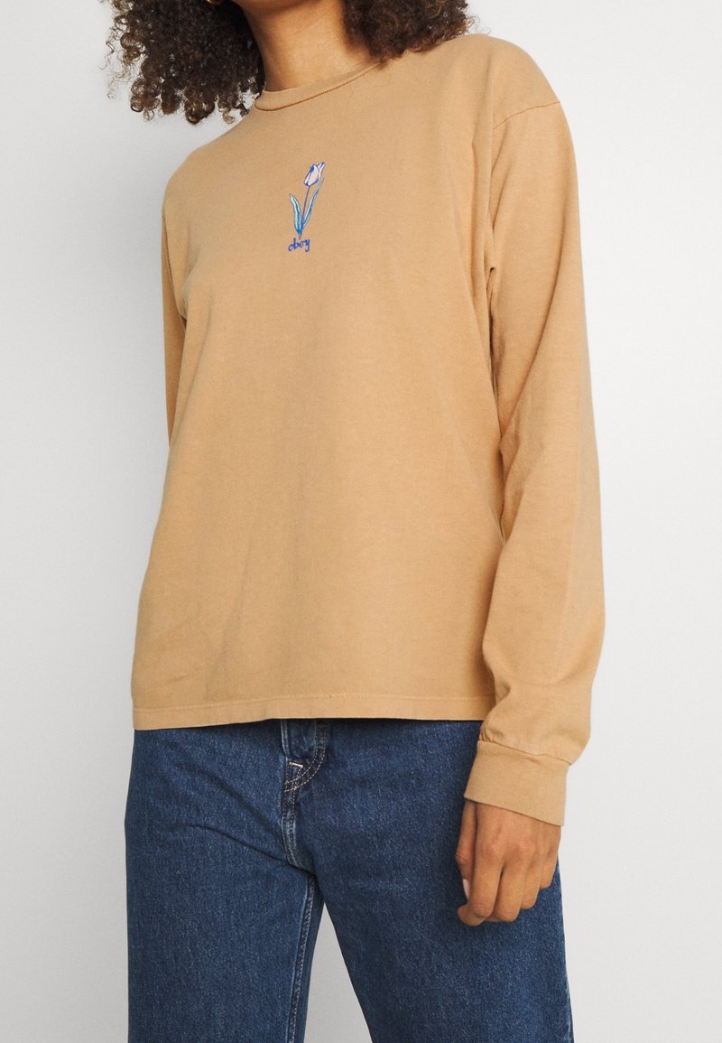 Långärmad, ljusbrun bomullsskjorta med rund hals, med broderad blommönster i blått och grönt i mitten.