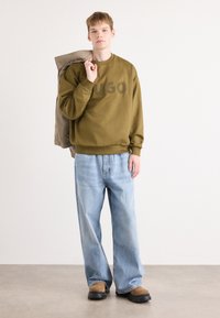Felpa verde oliva con logo "HUGO", abbinata a jeans baggy blu chiaro e scarpe slip-on marroni, mentre tiene una giacca marrone chiaro.