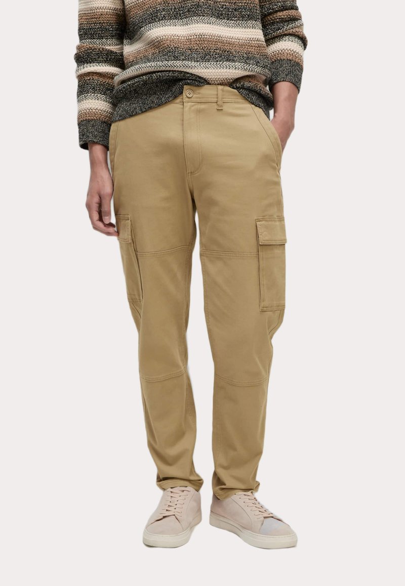 Beige cargo kalhoty s dvěma bočními kapsami, zapínáním na knoflík a uvolněným střihem, které jsou kombinovány se světlými teniskami a pruhovaným svetrem.