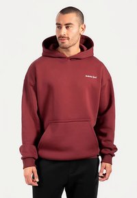 Felpa con cappuccio color bordeaux in tessuto spesso, con tasca frontale e polsini a coste. Presenta un piccolo logo bianco sul petto.