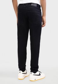 Blugi negri din denim cu croială strâmtă, buzunare în spate și o etichetă de piele pe talie, asortați cu adidași albi.