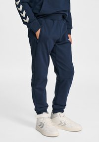 Mörkblåa träningsbyxor med blixtlåsförsedd ficka, elastisk midja och silverfärgade v-formade detaljer på övre vänstra benet. Matchas med vita sneakers.