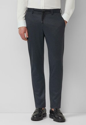 Man draagt donkergrijze, op maat gemaakte broek met zwarte riem, wit overhemd en zwarte leren schoenen, staand met één hand in de zak.