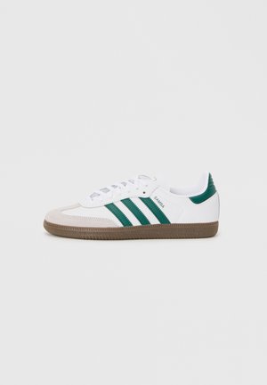 Weißer Sneaker mit grünen Streifen, Wildleder-Stopper und strukturiertem braunem Gummisohle. Verfügt über flache Schnürsenkel und einen gepolsterten Kragen.