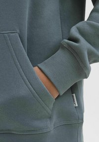 Sudadera gris con puños acanalados, bolsillo tipo canguro y una etiqueta que dice "ORIGINALS". Fabricada en suave algodón con una textura lisa.