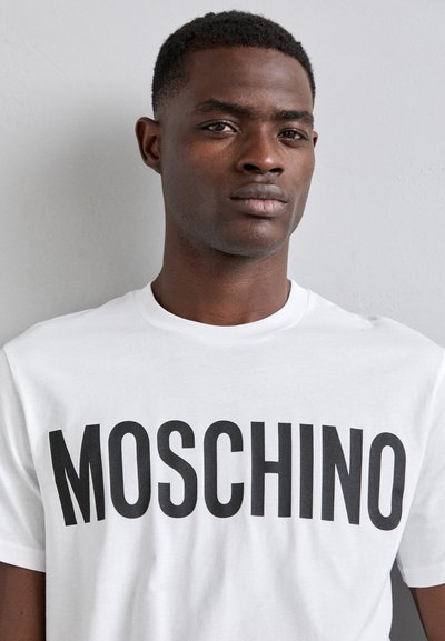 Balta medvilninė marškinėliai su ryškiu juodu "MOSCHINO" užrašu. Klasikinio apvalaus kaklo dizainas, trumpomis rankovėmis ir minkšta tekstūra.