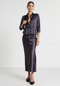 Frau trägt eine dunkelviolette Satinbluse mit Bindegürtel und passenden langen Rock, hält eine kleine schwarze Handtasche und trägt schwarze spitze Schuhe.