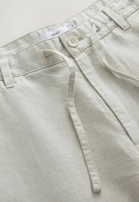 Pantalon en denim beige avec passants pour ceinture, un bouton blanc, des poches avant et une étiquette de marque visible "NEXT" à l'intérieur de la ceinture.