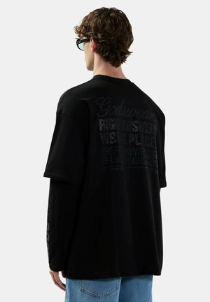 Giovane uomo con capelli ricci che indossa maglie nere a maniche lunghe e corte sovrapposte con testo sbiadito sul retro, jeans azzurri e occhiali da sole neri, di spalle.