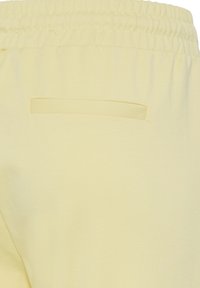 Section de pantalon jaune clair avec ceinture élastique et une poche passepoilée horizontale unique à l’arrière, tissu affichant une texture subtile.