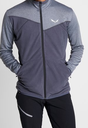 Homme portant une veste zippée texturée gris clair et gris foncé avec les mains dans les poches, associée à un pantalon noir avec une poche latérale zippée.